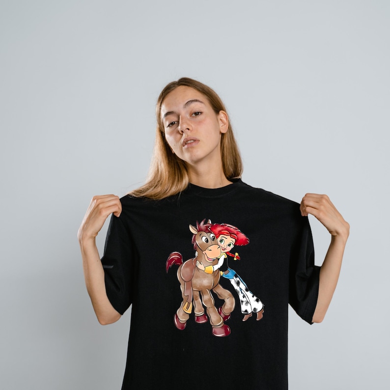 May include: Black t-shirt featuring a cartoon illustration of a girl with red hair hugging a brown horse. The girl is wearing a blue and white outfit. The horse has a yellow collar. The t-shirt is a solid black color.
