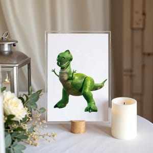 May include: A framed print of a green cartoon dinosaur with a friendly expression. The dinosaur is standing and waving. The frame is clear acrylic, supported by a small wooden block. A white candle and a lantern are also visible.