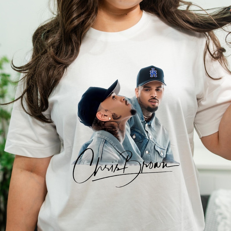 May include: White t-shirt featuring a graphic of Chris Brown in a denim jacket and a black cap. The shirt has the artist's signature in black cursive. The image is a portrait of the singer.