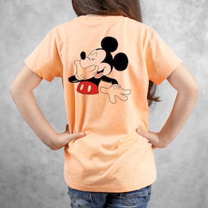Op de afbeelding: Perzikkleurig T-shirt met een afbeelding van Mickey Mouse die zijn mond bedekt en lacht. Het stripfiguur is in zwart, rood en wit. Het shirt heeft korte mouwen.