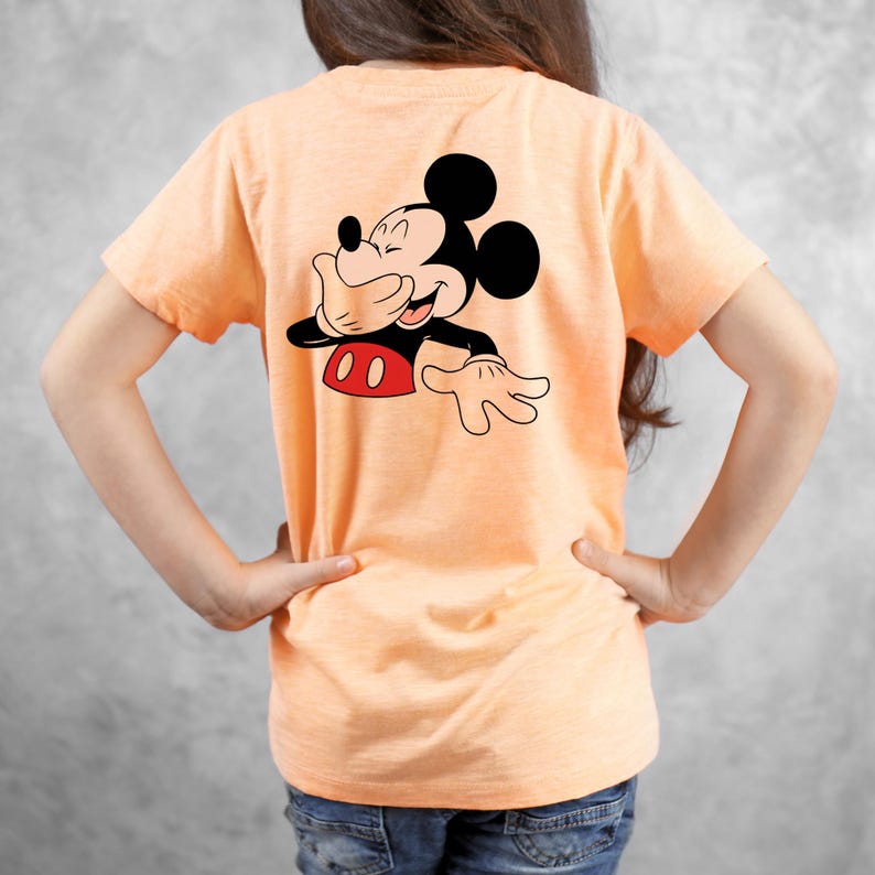 Op de afbeelding: Perzikkleurig T-shirt met een afbeelding van Mickey Mouse die zijn mond bedekt en lacht. Het stripfiguur is in zwart, rood en wit. Het shirt heeft korte mouwen.