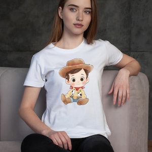 May include: White t-shirt featuring a cartoon illustration of a cowboy character. The cowboy is wearing a brown hat, a yellow shirt with black spots, blue jeans, and a red bandana. The design is centered on the shirt.