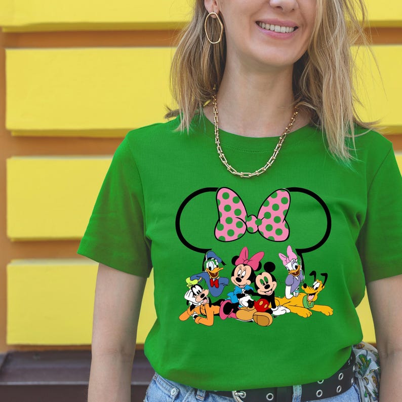 Op de afbeelding: Groen t-shirt met een grote Minnie Mouse strik met roze stippen en een zwarte omtrek. Het shirt heeft stripfiguren waaronder Mickey, Minnie, Donald, Daisy, Goofy en Pluto.