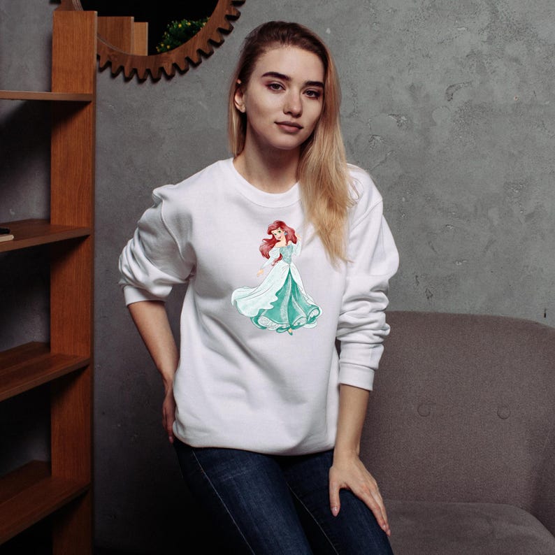 May include: White sweatshirt featuring a watercolor illustration of a princess in a teal and white gown with red hair. The sweatshirt is a crew neck style and is made of a soft material. The princess graphic is centered on the front.