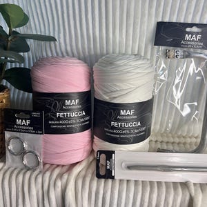 Può includere: Un assortimento di forniture per l'artigianato, tra cui due rocchetti di filato fettuccia rosa e bianco, anelli metallici, un manico per borsa in metallo e un uncinetto. I prodotti sono etichettati con il marchio "MAF Accessories".