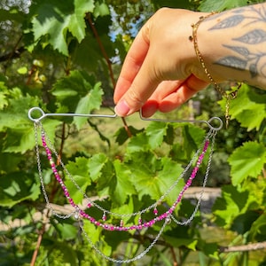 Könnte beinhalten: Ein mehrlagiges dekoratives Accessoire mit einem silberfarbenen Drahtrahmen und zwei Schlaufen an der Oberseite. Das Stück ist mit feinen silbernen Ketten und Strängen aus kleinen, leuchtend fuchsiafarbenen Perlen verziert, die einen gestuften Drapierungseffekt erzeugen.