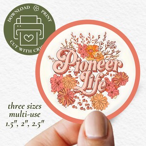 May include: A round sticker with a peach border and the words "Pioneer Life" in a retro font, surrounded by floral illustrations in shades of peach, orange, and pink. The sticker is held by a person's fingers. Additional text includes "three sizes multi-use 1.5", 2", 2.5" and "Download Print Cut with Cric".