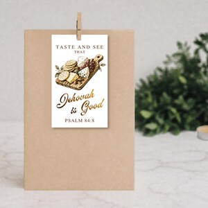May include: A rectangular white paper tag featuring a printed illustration of a wooden charcuterie board with cheese, crackers, and grapes. The text reads TASTE AND SEE THAT Jehovah is Good PSALM 84:8 in black and gold script lettering.