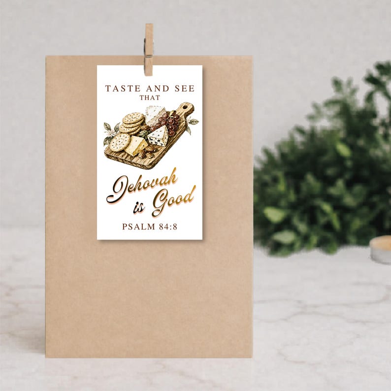 May include: A rectangular white paper tag featuring a printed illustration of a wooden charcuterie board with cheese, crackers, and grapes. The text reads TASTE AND SEE THAT Jehovah is Good PSALM 84:8 in black and gold script lettering.