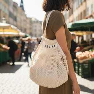 Può includere: Una borsa a rete in cotone color bianco sporco con una fascia in tessuto solido sulla parte superiore con il nome ELENA stampato in lettere maiuscole nere. La borsa è dotata di due tracolle e di una struttura flessibile a maglia aperta.