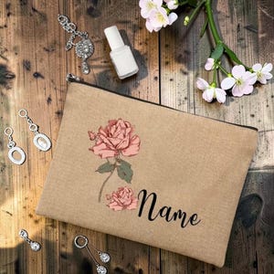 Op de afbeelding: Een beige canvas make-up tas met een zwarte rits, met een roze rozenillustratie en de naam "Name" in cursief. De tas wordt omringd door sieraden en een fles witte nagellak.
