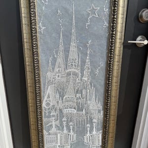 Könnte beinhalten: Ein gerahmtes Kunstwerk mit einer detaillierten weißen Illustration eines Schlosses mit mehreren Türmen und Türmchen, vor einem hellblauen Hintergrund. Der Rahmen ist goldfarben mit aufwendigen Details. Das Kunstwerk enthält sternförmige Verzierungen.