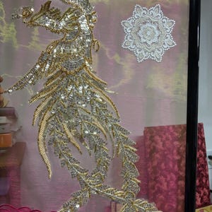Könnte beinhalten: Gerahmtes Kunstwerk mit einem detaillierten, verzierten Design einer vogelartigen Figur mit goldenen und silbernen Pailletten und Perlen. Ein weißes, sternförmiges Ornament ist ebenfalls vorhanden. Das Kunstwerk ist vor einem rosa Hintergrund mit einem bestickten Rand angeordnet.
