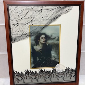 May include: Framed artwork featuring a portrait of a woman with dark hair, framed in a dark wood frame. The artwork is embellished with black lace and a decorative element. The portrait is set against a white background.