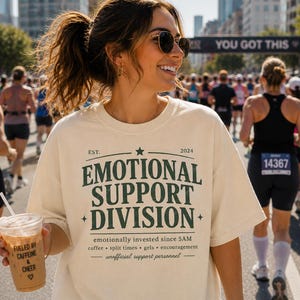Puede incluir: Camiseta color crema con el texto "EMOTIONAL SUPPORT DIVISION" en verde oscuro. La camiseta también tiene el texto "emotionally invested since 5AM" y "unofficial support personnel". Una mujer sostiene una taza de café con el texto "FUELED BY CAFFEINE & CHEER".
