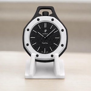 Peut inclure: Une montre Swatch Royal Pop noire et blanche sur un support blanc. La montre a un cadran noir avec des aiguilles et des marqueurs blancs. Le boîtier de la montre est noir avec une lunette blanche. Le texte "AP swatch" et "Royal Pop" est visible.