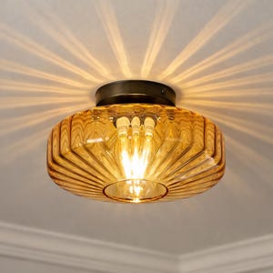 May include: A ceiling light with an amber glass shade featuring a ribbed design. The light fitting has a dark metal base and emits a warm glow. The design is reminiscent of vintage styles, suitable for home decor. The light's diameter is approximately 20 cm.