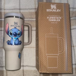 May include: A cream-colored Stanley Quencher H2.0 FlowState tumbler with a handle and a Stitch character graphic, accompanied by its brown cardboard packaging box labeled with the product name and a capacity of 40 oz and 1.18 L.
