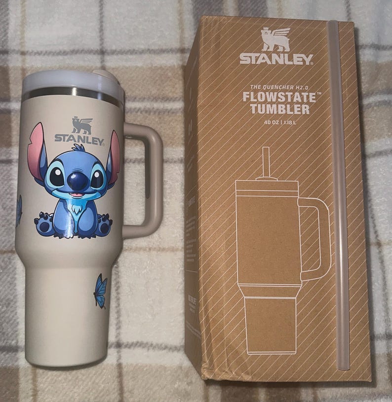 May include: A cream-colored Stanley Quencher H2.0 FlowState tumbler with a handle and a Stitch character graphic, accompanied by its brown cardboard packaging box labeled with the product name and a capacity of 40 oz and 1.18 L.