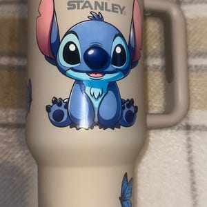 May include: A beige Stanley tumbler with a handle and a white lid. The front features a graphic of the blue character Stitch with large ears, sitting down. A small blue butterfly is printed on the lower side of the cup below the character.