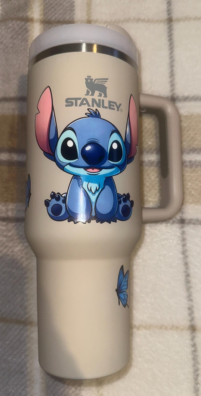 May include: A beige Stanley tumbler with a handle and a white lid. The front features a graphic of the blue character Stitch with large ears, sitting down. A small blue butterfly is printed on the lower side of the cup below the character.