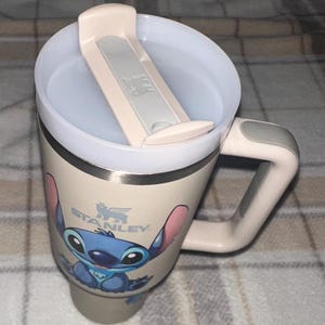 May include: A cream-colored Stanley travel mug featuring a graphic of the character Stitch on the front. The mug has a matching cream-colored handle with a gray accent, a clear plastic lid with a cream-colored sliding closure, and a stainless steel rim.