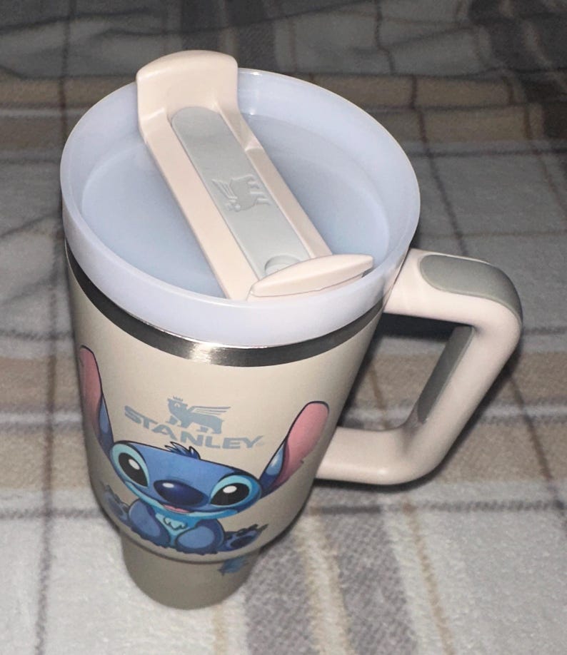 May include: A cream-colored Stanley travel mug featuring a graphic of the character Stitch on the front. The mug has a matching cream-colored handle with a gray accent, a clear plastic lid with a cream-colored sliding closure, and a stainless steel rim.