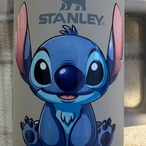 May include: A light beige Stanley brand tumbler featuring a graphic of the blue character Stitch from Lilo and Stitch. The character has large ears with pink inner sections, a dark blue nose, and is sitting in a forward-facing position.