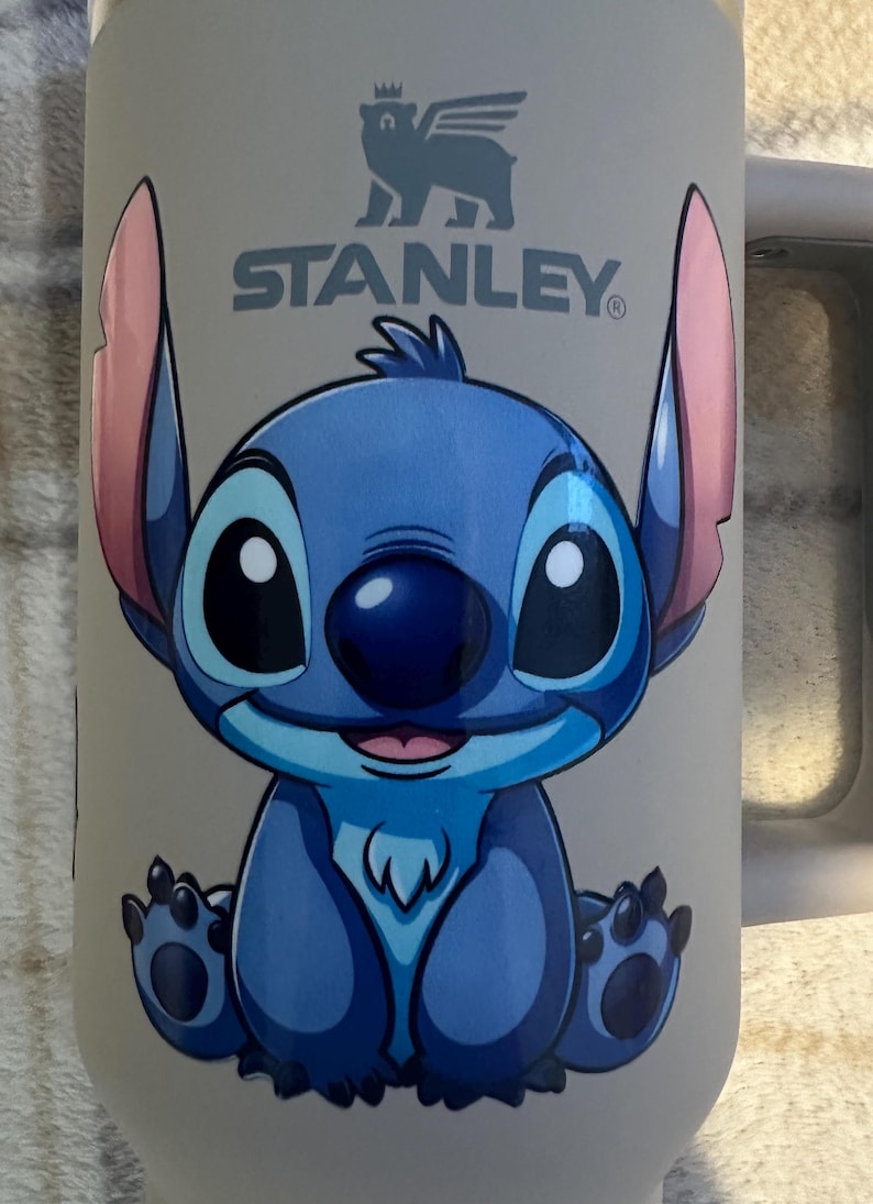 May include: A light beige Stanley brand tumbler featuring a graphic of the blue character Stitch from Lilo and Stitch. The character has large ears with pink inner sections, a dark blue nose, and is sitting in a forward-facing position.