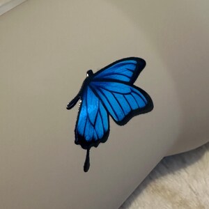 May include: A vibrant blue butterfly decal with distinct black outlines and a singular black teardrop-shaped accent extending from the lower wing. The butterfly graphic features deep blue shading within the wings and is applied to a smooth surface.