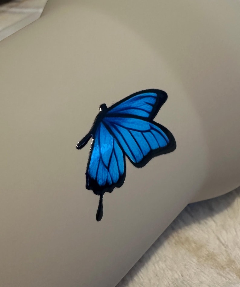 May include: A vibrant blue butterfly decal with distinct black outlines and a singular black teardrop-shaped accent extending from the lower wing. The butterfly graphic features deep blue shading within the wings and is applied to a smooth surface.