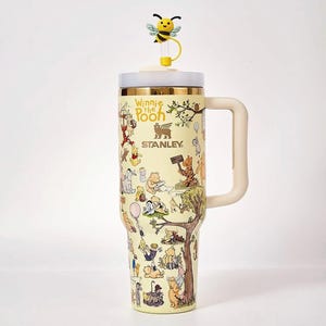 Könnte beinhalten: Ein blassgelber Stanley-Becher mit Winnie Puuh-Motiven, einem seitlichen Griff und einem transparenten Deckel mit gelbem Bienen-Strohhalm-Aufsatz. Der Becher zeigt verschiedene Illustrationen von Puuh und seinen Freunden im Wald.