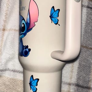 May include: A cream-colored insulated tumbler with a handle, featuring a graphic of the blue character Stitch and two bright blue butterflies. The tumbler has a metallic rim and a light-colored lid. The Stanley brand logo is visible near the top.