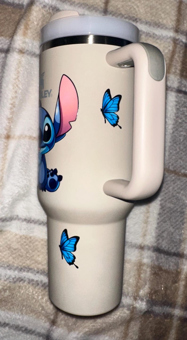 May include: A cream-colored insulated tumbler with a handle, featuring a graphic of the blue character Stitch and two bright blue butterflies. The tumbler has a metallic rim and a light-colored lid. The Stanley brand logo is visible near the top.