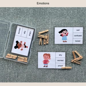 May include: Educational emotion flashcards with illustrations and wooden clothespins. The cards feature words like "worried," "silly," and "angry." The set includes a clear plastic container and the word "Emotions" at the top.