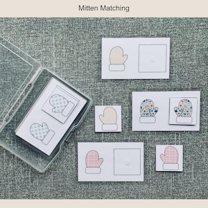 May include: A clear plastic box containing mitten-themed matching cards. Several white cards with mitten illustrations are displayed, including patterned and floral designs. The text "Mitten Matching" is at the top.
