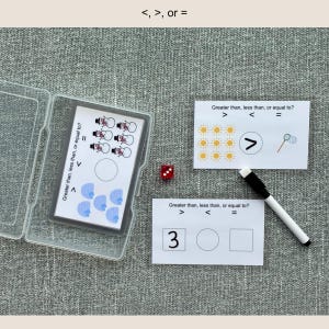 May include: Educational game with cards and a dry-erase marker. The cards feature the phrases "Greater than, less than, or equal to?" and symbols. The game includes a clear plastic case, a die, and cards with suns, snowmen, and numbers.