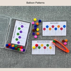 May include: A balloon pattern matching educational activity set featuring three white cards with printed balloon sequences, a clear plastic storage case, a collection of small multicolored pom-poms, and a pair of orange plastic tweezers.