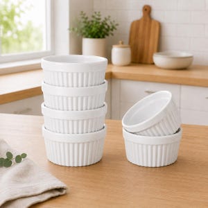May include: A set of five white ceramic ramekins with a fluted exterior design. Four of the ramekins are stacked vertically in a single column, while one ramekin is positioned to the right, resting on its side against the base of the stack.