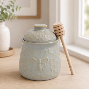 May include: A light blue ceramic honey pot with a matching lid, featuring an embossed honeycomb pattern and a bee motif on the front. A light-colored wooden honey dipper rests against the side of the pot, which has a speckled finish.