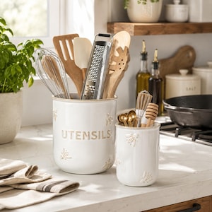 May include: Two white ceramic crocks with embossed bee motifs and tan rims. The larger crock is labeled UTENSILS and holds a metal grater, wooden spatula, wooden spoon, and wire whisk. The smaller crock contains a honey dipper and small spoons.