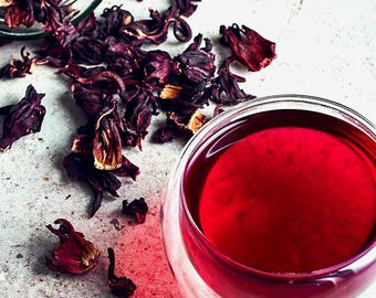 May include: A glass of vibrant red tea next to a spilled jar of dried hibiscus flowers. The tea is a deep crimson color, and the dried flowers are a dark maroon. The image is set against a light gray background.