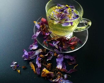 May include: A clear glass teacup and saucer filled with a light green liquid and purple flower petals. The cup sits on a clear saucer, with scattered petals around it. The background is a dark surface.
