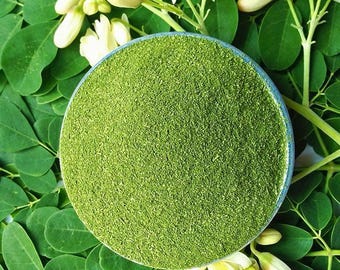 May include: A top-down view of a bowl filled with finely ground, bright green powder, likely a dietary supplement. The bowl is surrounded by fresh green leaves and delicate white and yellow flowers, creating a natural, organic aesthetic.
