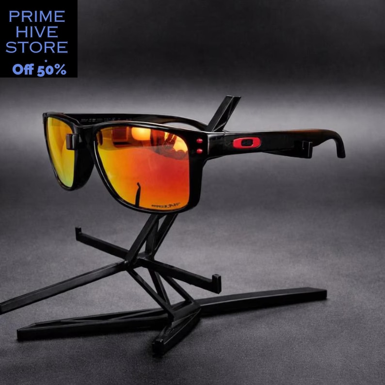 Peut inclure: Lunettes de soleil noires avec des verres orange, pr&eacute;sent&eacute;es sur un pr&eacute;sentoir noir. Les lunettes ont une monture rectangulaire avec des d&eacute;tails rouges. Le texte "PRIME HIVE STORE" et "Off 50%" est en haut &agrave; gauche.