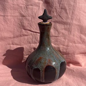 May include: A ceramic bottle with a rounded base and a narrow neck, featuring a glossy glaze that transitions from a deep black at the bottom to a mottled blue and orange drip pattern. A dark, cross-shaped stopper sits atop the bottle opening.