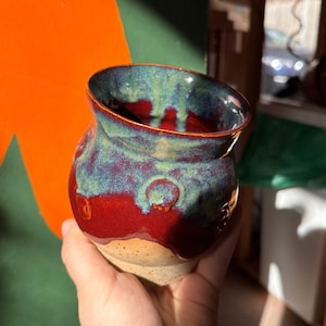 May include: A handmade ceramic vase with a unique design. The vase features a red base, transitioning to a blue-green upper section. The rim is slightly flared. The vase is held in a hand, showcasing its size and shape.