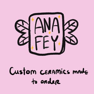 May include: A graphic logo on a light pink background featuring the text ANA FEY inside a rectangular shape with black outlines and stylised wings on both sides. Small yellow stars surround the text. Below, the text reads Custom ceramics made to order.