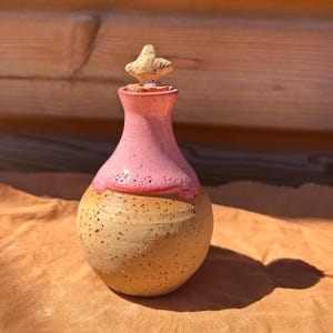 May include: A handmade ceramic bottle with a pink neck and a speckled tan body. The bottle is topped with a tan stopper. The bottle is sitting on a tan surface.