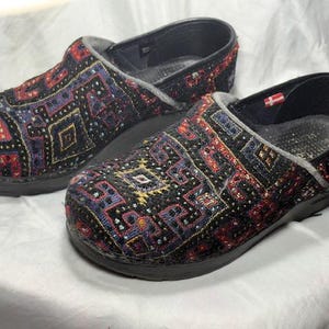 May include: Pair of black clogs with a colorful, patterned textile upper. The pattern includes red, blue, and gold geometric shapes. The clogs have a black sole and a gray trim.